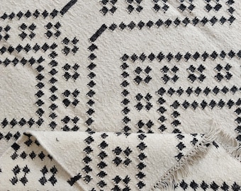 May include: Cream-colored woven rug with a geometric pattern in black. The design features squares and diamond shapes. The rug has a rolled-up section, revealing a fringed edge. The rug is made of a textured material.