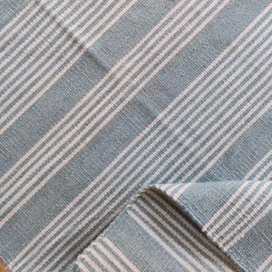 Pu&ograve; includere: Un tessuto a righe blu e bianche, possibilmente un tappeto o una coperta, con un motivo diagonale. Il tessuto ha un aspetto strutturato, con le strisce bianche che contrastano con lo sfondo blu. Il tessuto &egrave; piegato, rivelando il rovescio.