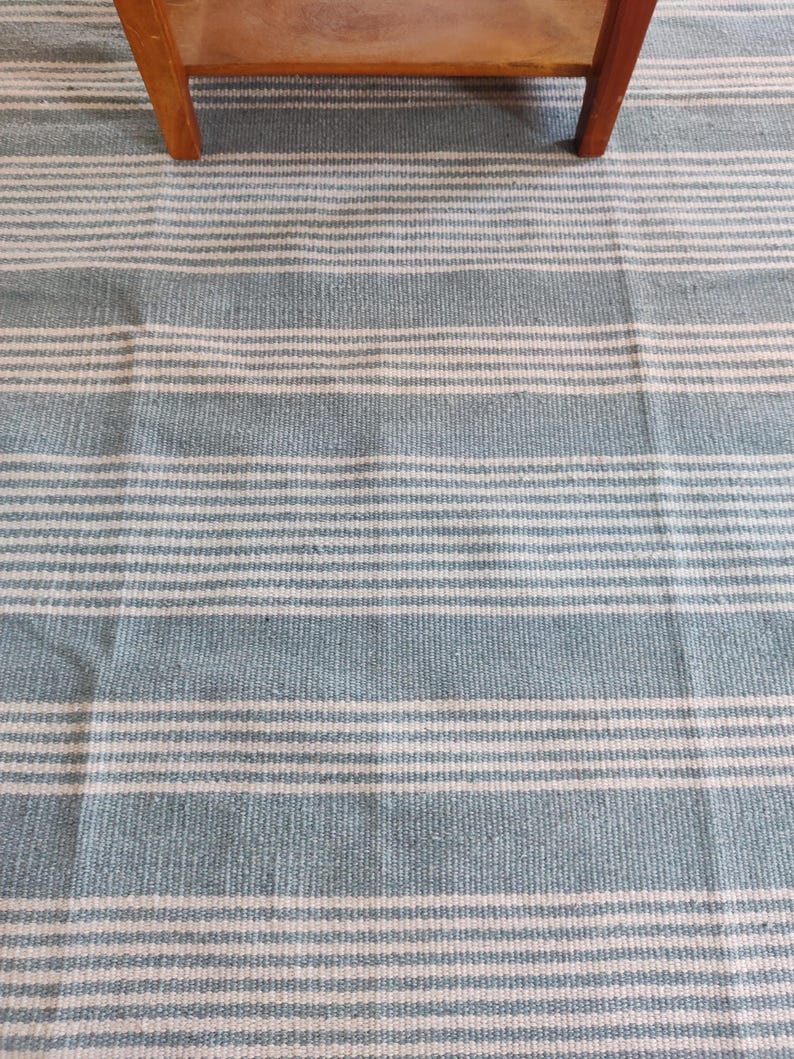 Pu&ograve; includere: Un tessuto a righe blu e bianche, possibilmente un tappeto o una coperta, con un motivo orizzontale ripetuto. Il tessuto &egrave; di colore azzurro con strisce bianche. Una gamba di tavolo in legno &egrave; visibile nella parte superiore dell'immagine.