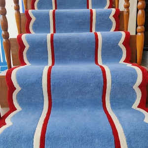 May include: A blue stair runner with a scalloped red border and a central white stripe. The runner is installed on wooden stairs with wooden banisters. The runner's design adds a pop of color and visual interest to the staircase.