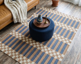 Op de afbeelding: Een rechthoekig vloerkleed met een herhalend patroon van blauwe en lichtbruine verticale strepen, afgewisseld met een wit en lichtbruin geblokte rand. Het kleed ligt op een houten vloer onder een ronde marineblauwe poef met een houten dienblad.