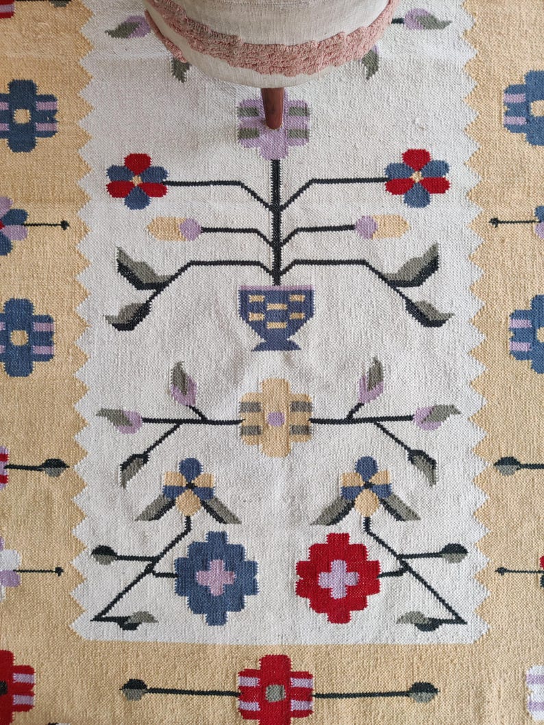 Puede incluir: Una alfombra tejida que presenta un panel central blanco con un patr&oacute;n floral sim&eacute;trico en rojo, azul, lavanda y gris. El dise&ntilde;o incluye flores estilizadas, hojas y un motivo de jarr&oacute;n central, enmarcado por un borde dentado y paneles laterales de color tostado.