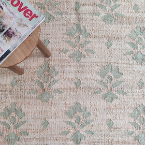 Puede incluir: Una alfombra de yute de color crema con un patrón floral verde claro. La alfombra es texturizada y tiene un brillo sutil. Una revista con la palabra "Cover" en la portada está sentada en un taburete de madera sobre la alfombra.