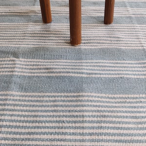 Pu&ograve; includere: Primo piano di un tappeto a righe blu e bianche con tre gambe in legno marrone. Il tappeto ha un aspetto strutturato con strisce orizzontali. Le gambe in legno sono cilindriche e sembrano far parte di un tavolino o sgabello.