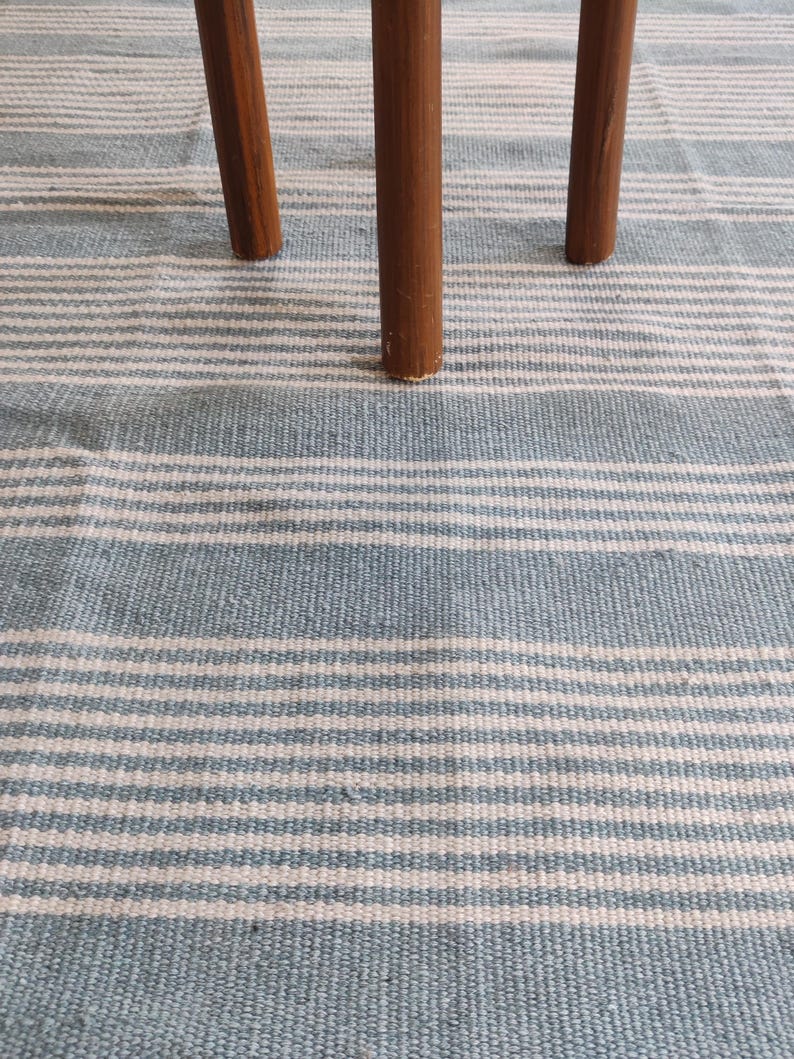 Pu&ograve; includere: Primo piano di un tappeto a righe blu e bianche con tre gambe in legno marrone. Il tappeto ha un aspetto strutturato con strisce orizzontali. Le gambe in legno sono cilindriche e sembrano far parte di un tavolino o sgabello.