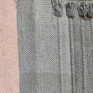 May include: Close-up of a woven rug with a light pink stripe and a dark gray stripe. The rug has a fringe on the right side.
