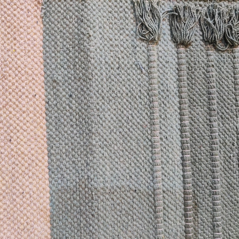 May include: Close-up of a woven rug with a light pink stripe and a dark gray stripe. The rug has a fringe on the right side.