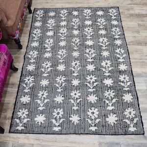 Puede incluir: Una alfombra rectangular con estampado en blanco y negro. La alfombra presenta un diseño floral repetitivo en blanco sobre un fondo negro. La alfombra está colocada sobre un suelo de madera, con una silla marrón visible en el fondo.
