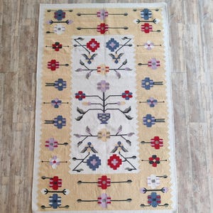 Puede incluir: Una alfombra rectangular que presenta un panel central blanco con un motivo floral sim&eacute;trico en rojo, azul, morado y amarillo. El borde es de un color beige texturizado con patrones de flores geom&eacute;tricas repetitivas en rojo, azul y blanco.