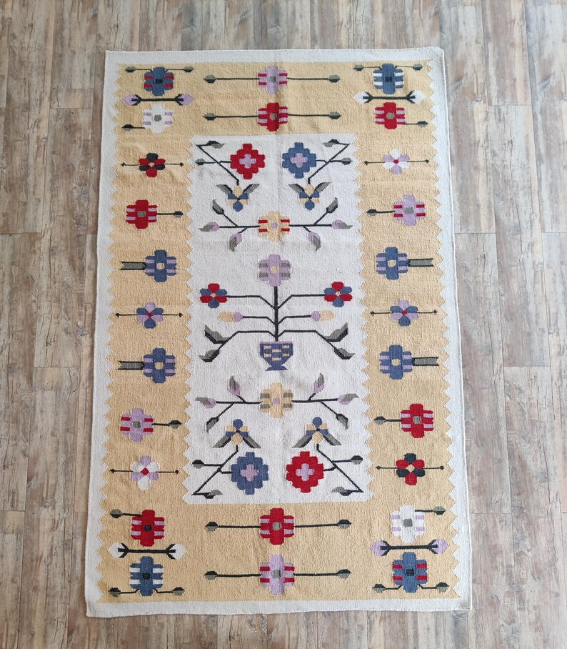 Puede incluir: Una alfombra rectangular que presenta un panel central blanco con un motivo floral sim&eacute;trico en rojo, azul, morado y amarillo. El borde es de un color beige texturizado con patrones de flores geom&eacute;tricas repetitivas en rojo, azul y blanco.