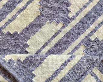 May include: A woven rug with a geometric pattern. The rug features alternating diagonal stripes of white and periwinkle blue. The edges of the rug are finished with a periwinkle blue border. The rug is made of a textured material.