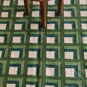 Puede incluir: Una alfombra de patr&oacute;n geom&eacute;trico verde y blanco con un taburete de madera en la parte superior. El taburete tiene una revista en la parte superior con el t&iacute;tulo "Awaken the Spirits".