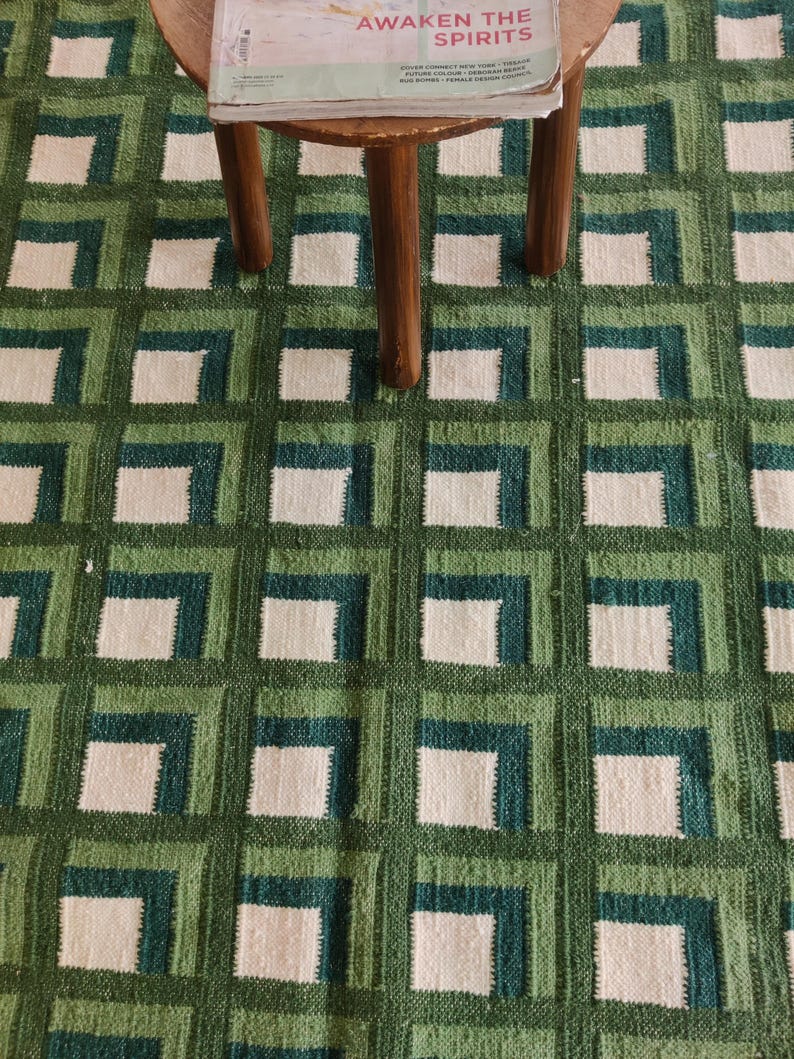 Puede incluir: Una alfombra de patr&oacute;n geom&eacute;trico verde y blanco con un taburete de madera en la parte superior. El taburete tiene una revista en la parte superior con el t&iacute;tulo "Awaken the Spirits".