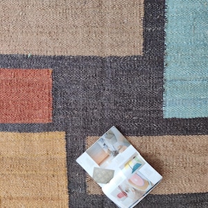 May include: A flat woven rug with a geometric pattern in shades of brown, orange, blue, and black. A magazine is lying open on the rug.