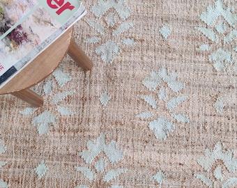 May include: A light brown rug with a white and light blue geometric pattern. The rug has a woven texture and is made of natural fibers.
