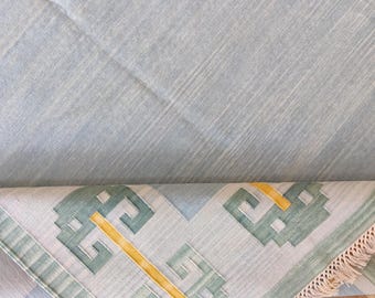Puede incluir: Una alfombra azul claro con un patrón geométrico en tonos verdes y amarillos. La alfombra tiene flecos en un borde y está parcialmente enrollada, revelando el patrón. La alfombra está hecha de un material tejido.