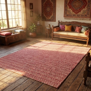 May include: A sunlit interior with wooden furniture, including a bench, chairs, and a trunk. A large red rug covers the floor. Decorative rugs hang on the walls. Plants and pillows add color to the room. A window lets in natural light.