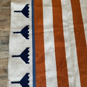 May include: A white and orange striped rug with a blue border. The border has a repeating pattern of dark blue stylized flowers.