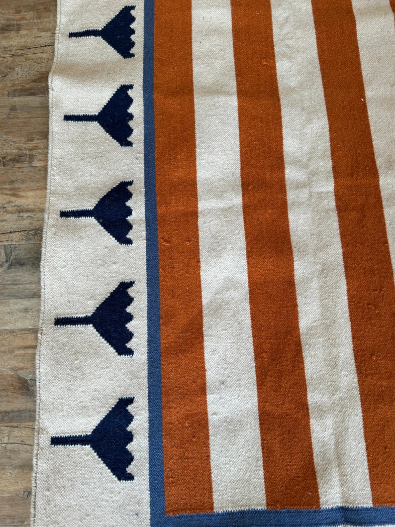 May include: A white and orange striped rug with a blue border. The border has a repeating pattern of dark blue stylized flowers.