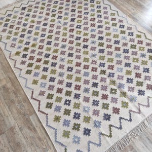 May include: A white rug with a geometric pattern of small squares in shades of green, blue, purple, and brown. The rug has a fringe border.