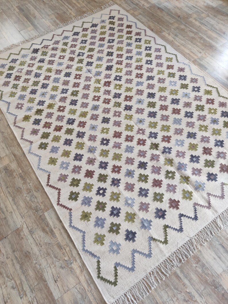 May include: A white rug with a geometric pattern of small squares in shades of green, blue, purple, and brown. The rug has a fringe border.