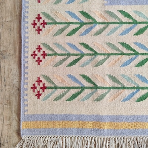 Könnte beinhalten: Ein gewebter Teppich mit cremefarbener Basis und einem sich wiederholenden Muster aus horizontalen Blumenstielen in Grün, Blau und Pfirsich, akzentuiert durch rote Blüten im Kreuzstich-Stil. Der Teppich hat einen hellvioletten Rand, einen gelben Streifen und weiße Fransen.