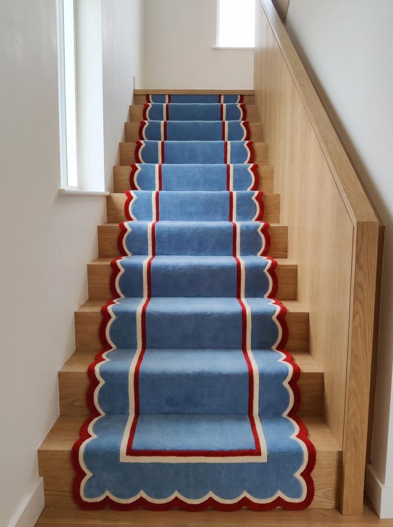 May include: A staircase with light blue carpet runners. The runners have a scalloped edge with a red border and a white inner stripe. The stairs are made of light-colored wood.