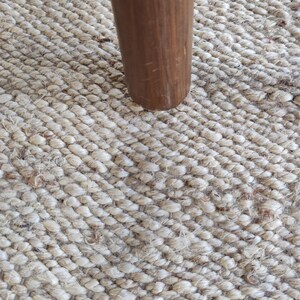 K&ouml;nnte beinhalten: Nahaufnahme eines Tischbeins aus Holz, das auf einem Naturfaser-Teppich ruht. Das Bein ist in einem satten Braun gehalten und hat eine glatte, leicht strukturierte Oberfl&auml;che. Der Teppich besteht aus dicht gewebten Fasern und hat eine hellbeige Farbe.
