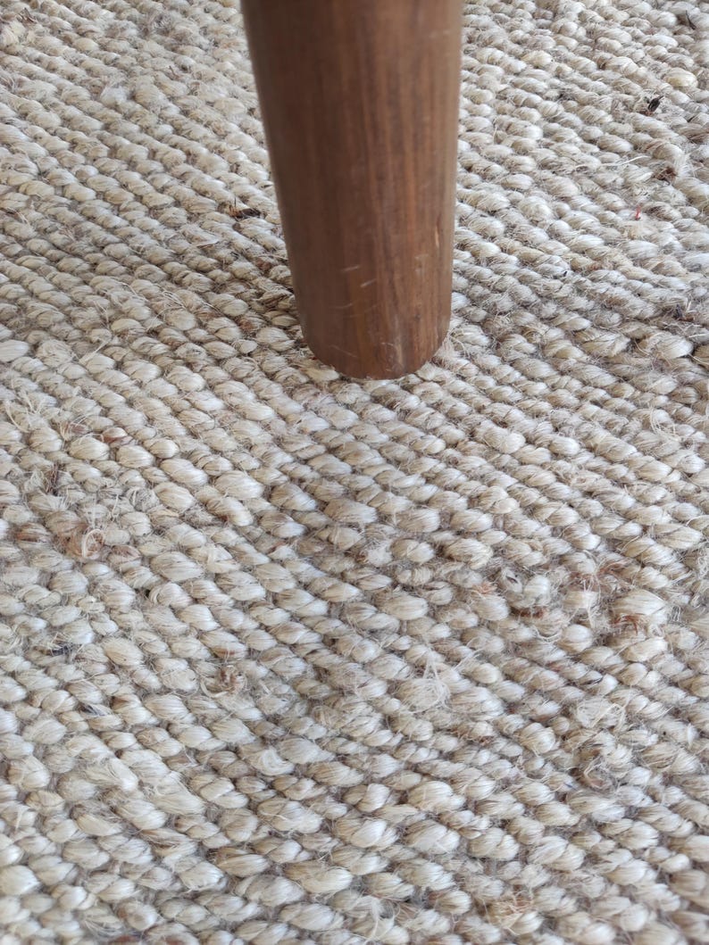 K&ouml;nnte beinhalten: Nahaufnahme eines Tischbeins aus Holz, das auf einem Naturfaser-Teppich ruht. Das Bein ist in einem satten Braun gehalten und hat eine glatte, leicht strukturierte Oberfl&auml;che. Der Teppich besteht aus dicht gewebten Fasern und hat eine hellbeige Farbe.