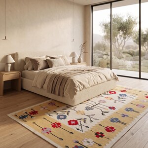 Puede incluir: Una alfombra rectangular que presenta un centro de color crema con un patr&oacute;n floral repetitivo en rojo, azul y morado claro. La alfombra tiene bordes anchos de color amarillo mostaza en los extremos superior e inferior, decorados con motivos florales.