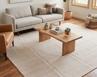 May include: A large rectangular area rug featuring a light beige colour and a subtle, repeating wavy herringbone texture. The rug is positioned on a wooden floor, covering a significant portion of the space beneath a wooden coffee table and sofa.