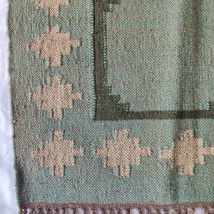 May include: A woven rug with a geometric pattern in shades of green and beige. The rug has a fringe border.