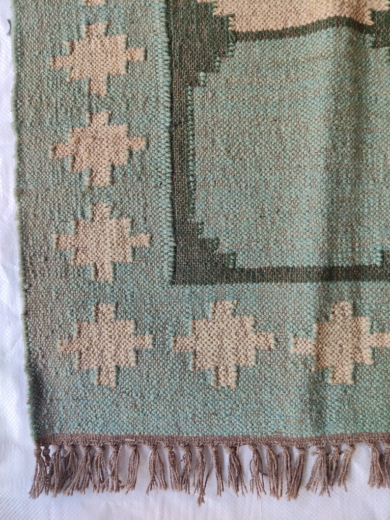 May include: A woven rug with a geometric pattern in shades of green and beige. The rug has a fringe border.