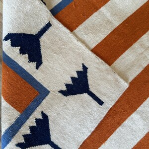 May include: A white rug with orange and blue stripes and a geometric pattern of dark blue stylized leaves.