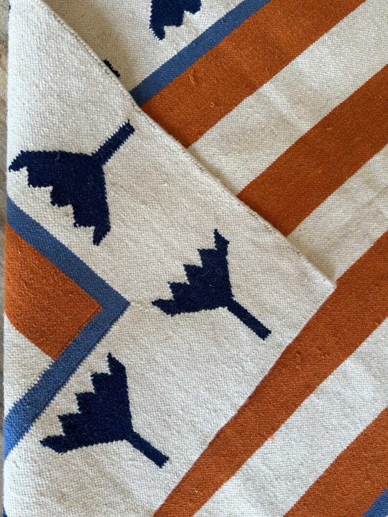May include: A white rug with orange and blue stripes and a geometric pattern of dark blue stylized leaves.