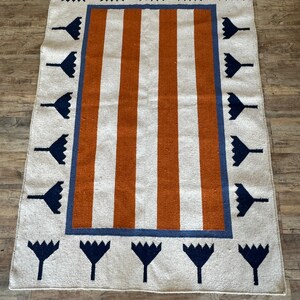 May include: A white and orange striped rug with a blue and white border. The border features a repeating pattern of stylized flowers.
