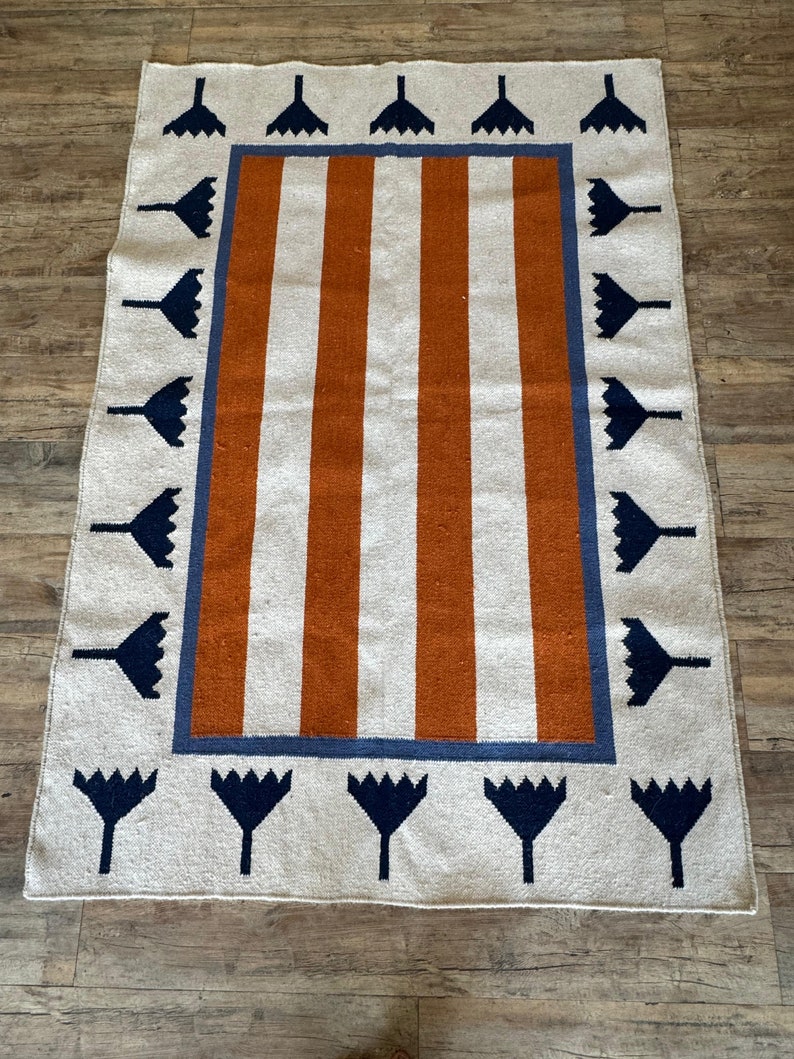 May include: A white and orange striped rug with a blue and white border. The border features a repeating pattern of stylized flowers.