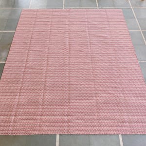 May include: A large, rectangular rug with a woven texture. The rug features a pattern of alternating red and pink horizontal stripes. The rug is placed on a tiled floor, and the overall aesthetic is simple and functional.