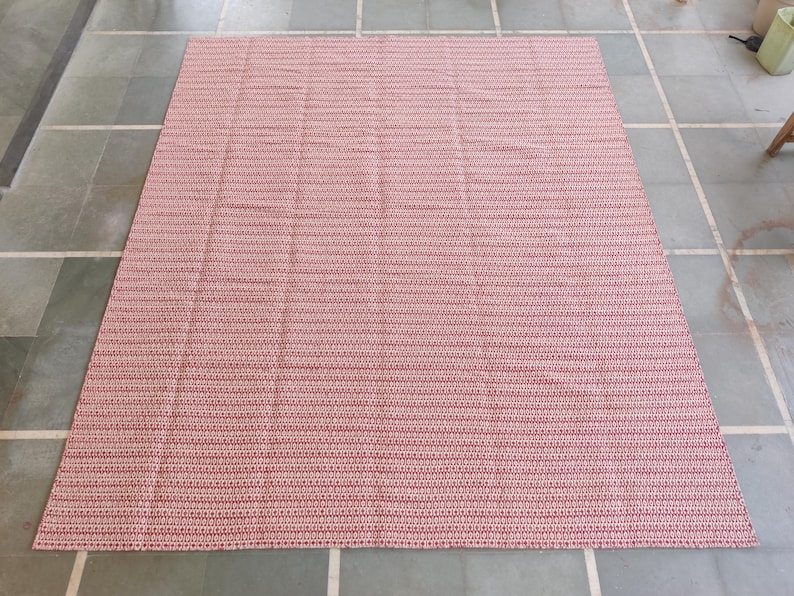 May include: A large, rectangular rug with a woven texture. The rug features a pattern of alternating red and pink horizontal stripes. The rug is placed on a tiled floor, and the overall aesthetic is simple and functional.