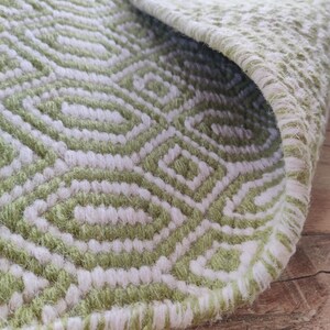 May include: Close-up of a green and white woven rug with a geometric pattern. The rug has a fringed edge and is made of soft, fluffy fibers.