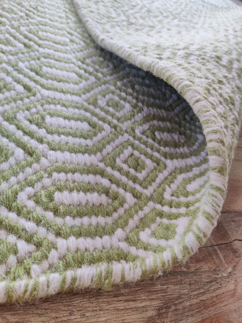 May include: Close-up of a green and white woven rug with a geometric pattern. The rug has a fringed edge and is made of soft, fluffy fibers.