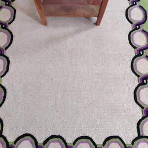 May include: A rectangular area rug with a light beige center and a patterned border. The border features a chain-like design in green, purple, black, and off-white. A wooden table with a magazine on top is partially visible.