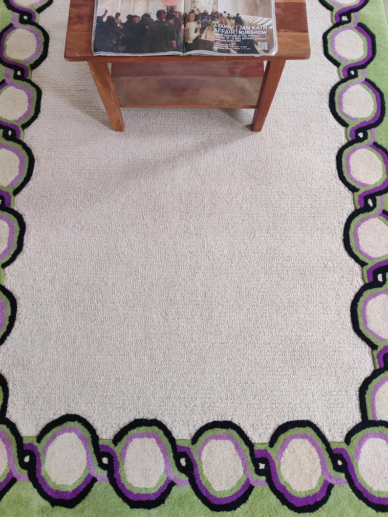 May include: A rectangular area rug with a light beige center and a patterned border. The border features a chain-like design in green, purple, black, and off-white. A wooden table with a magazine on top is partially visible.