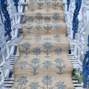 May include: A beige and blue patterned jute rug runner with a floral design. The rug is laid on a set of white painted stairs with blue metal railings.