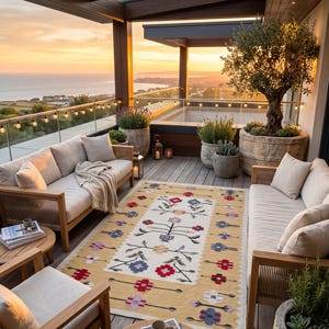 Puede incluir: Una alfombra rectangular con un centro de color crema que presenta un patr&oacute;n floral sim&eacute;trico en rojo, azul y morado, bordeada por una ancha franja amarilla con motivos florales repetitivos, colocada sobre una terraza de madera entre dos sof&aacute;s beige.