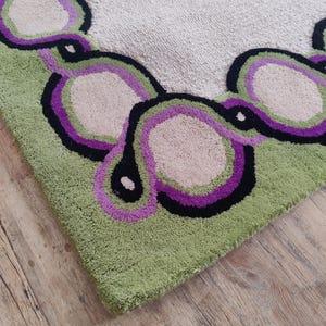 May include: Close-up of a green, beige, black, and purple rug. The rug has a repeating pattern of connected, abstract shapes in green, purple, black, and beige. The rug is on a wooden floor.
