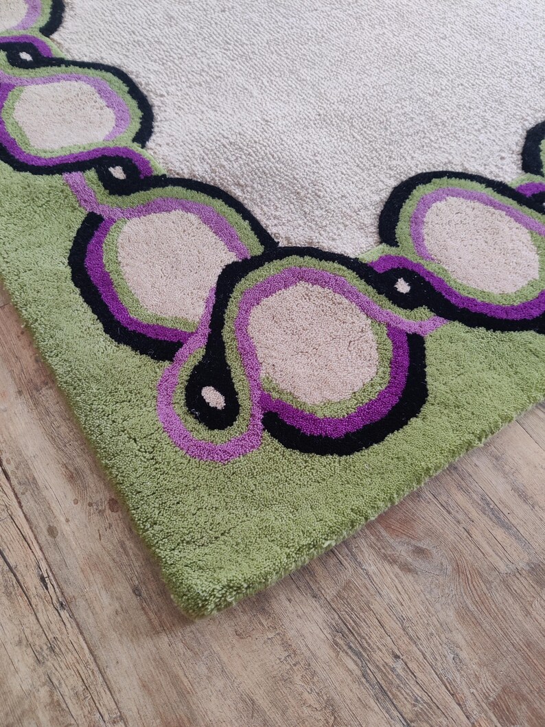 May include: Close-up of a green, beige, black, and purple rug. The rug has a repeating pattern of connected, abstract shapes in green, purple, black, and beige. The rug is on a wooden floor.