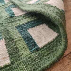 Puede incluir: Primer plano de una alfombra tejida verde y blanca con un patr&oacute;n geom&eacute;trico. La alfombra tiene un borde de flecos y est&aacute; hecha de un material suave y texturizado.