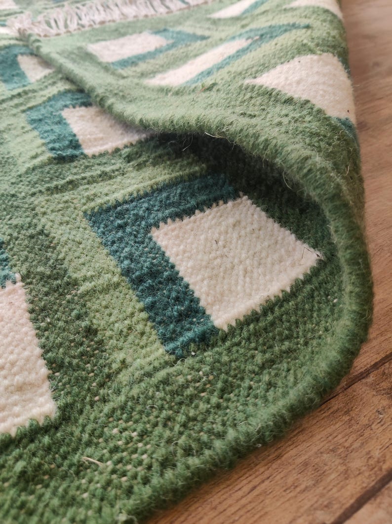 Puede incluir: Primer plano de una alfombra tejida verde y blanca con un patr&oacute;n geom&eacute;trico. La alfombra tiene un borde de flecos y est&aacute; hecha de un material suave y texturizado.