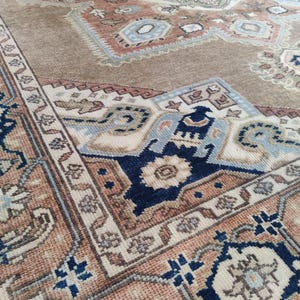 May include: A close-up of a beige rug with a detailed pattern in shades of blue, brown, and cream. The rug has a central medallion with a floral design and a border with paisley motifs.